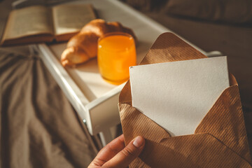 Close-up of a hand holding a kraft envelope with a blank card, with a breakfast tray in the...