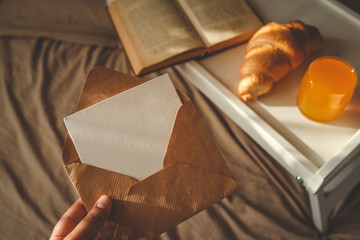 Close-up of a hand holding a kraft envelope with a blank card, with a breakfast tray in the...