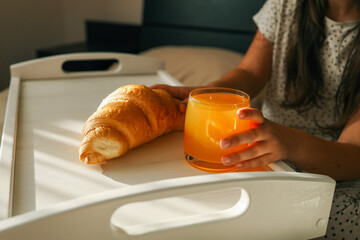 Child's hands reaching for a croissant and orange juice on a breakfast tray in bed, soft sunlight...