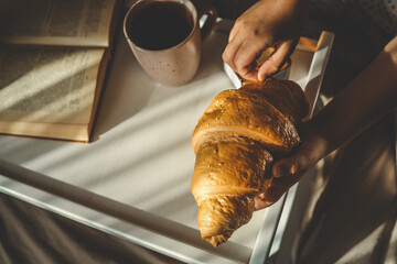 Cozy morning scene with a child holding a croissant in bed, next to a tray with coffee, candle,...