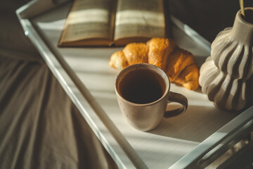 Beautiful morning scene with a croissant, candle, coffee, open book, and decorative vase on a white tray in bed, softly lit by natural sunlight through curtains
