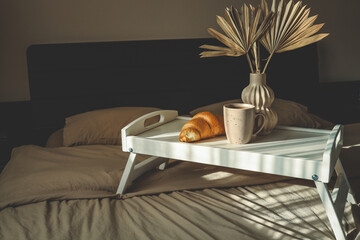 Minimal breakfast tray with a cup of coffee, croissant, and decorative vase on a cozy bed,...