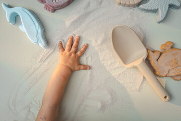 Top view of a child’s hand playing with sand, surrounded by pastel-colored sea creature toys and a beige scoop. The hand leaves playful marks in the sand, evoking sensory exploration and summer joy