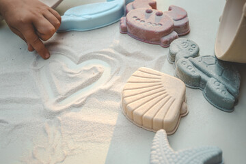 Child’s hand drawing a heart in fine white sand, surrounded by pastel sea-themed toys including a...
