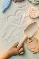 Child’s hand drawing a heart in fine white sand, surrounded by pastel sea-themed toys including a crab, dolphin, and shell. A sweet and playful moment of creativity and tactile exploration