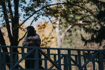 A loving couple embraces on a wooden bridge amidst a lush forest, capturing a moment of affection and tranquility. Sunlight filters through the trees, creating a romantic atmosphere.
