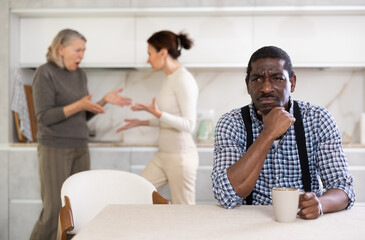Obraz premium Sad middle-aged man sitting at the kitchen table with his back to old and middle-aged women quarreling together