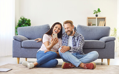 Obraz premium Young couple sitting on floor in living room at home using mobile phone for internet and social media or having online video call. Happy man and woman watching funny video on smartphone.