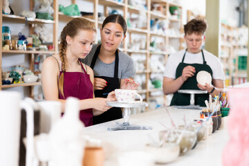 Young female teacher teaching teenage boy and girl students how to paint ceramic cup in ceramic workshop