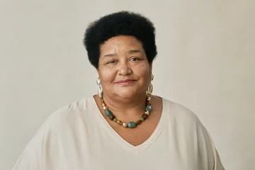 Fotobehang Portrait of mature African American woman with short curly hair wearing beaded necklace and white top, smiling softly at camera in studio © Seventyfour