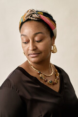 Vertical shot of Black woman wearing colorful headscarf and matching jewelry looking down with contemplative expression while posing in studio against beige background