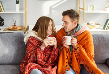 Young couple at home, sitting covered with warm blanket on sofa with hot drink, suffer with fever,...