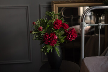 A bouquet of vibrant red peonies in a black vase, placed on a table in a stylish interior. Soft lighting enhances the springtime ambiance with candles nearby.