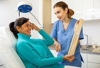 Skilled female beautician giving consultation to woman patient with mirror in aesthetic clinic