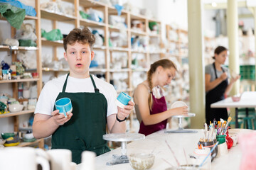 Young man in an apron demonstrates clay mugs, which he himself painted with colored paints