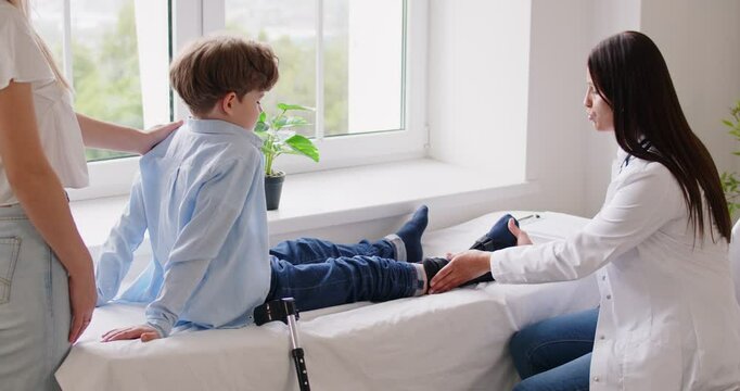 Female traumatologist talking about treatment and examining little kid patient with injured leg sitting on hospital bed. Boy in orthopedic bandage is supported by his mother during medical inspection.