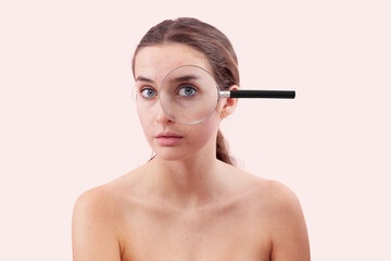 Obraz premium Woman portrait with no makeup, her eye examined under a magnifying glass. Authentic female face with real skin, acne, and blemishes before skincare or beauty treatment, isolated on a pink background