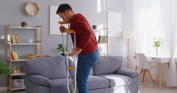Man with trauma, broken leg, man wearing foot support with walking stick crutches, patient wearing sling support after accident, traumatic stress, accident, sitting at home on sofa, resting to relax