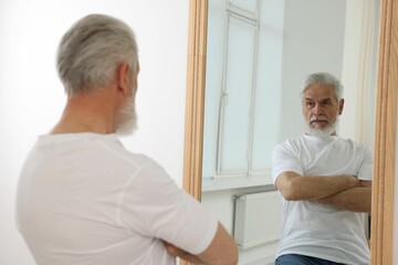 Handsome senior man looking at himself in mirror indoors