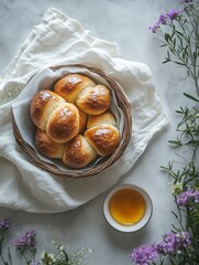 Fresh golden bread rolls in rustic basket on white linen with honey dip and spring flowers – cozy farmhouse breakfast or brunch flat lay