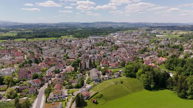 Aerial flyover historic town of Hinwil in Switzerland. Church tower and houses in center of city. Wide shot. Sunny day with green picturesque landscape in distance. Clouds at sky in spring.