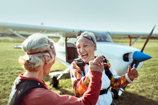 A senior lesbian couple stands on a field, airplane in the backdrop, beaming with pride and exhilaration after skydiving. Their smiles hint at a bucket list dream fulfilled.