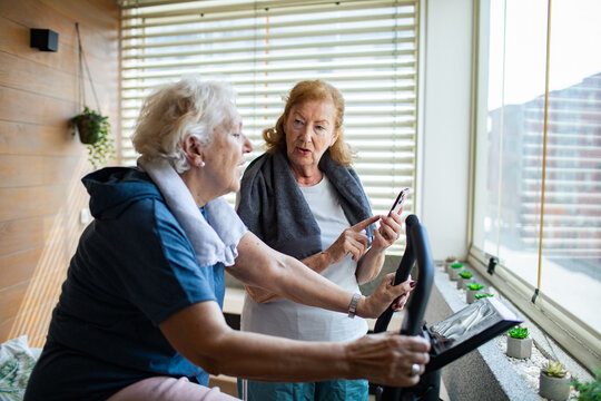 Senior lesbian couple using exercise bike and fitness app together at home gym - Powered by Adobe