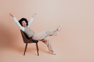 Confident young businesswoman in stylish beige attire celebrates success on wooden chair against...