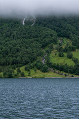 Waterfalls cascade down a lush green hillside into Storfjorden, Norway, with the calm waters of the fjord in the foreground and low clouds covering the mountain tops