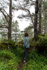 Young woman seen from behind exploring a lush Nordic forest, dressed in hiking gear and holding a camera, surrounded by mossy rocks, pines, and natural silence