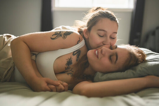 Happy couple cuddling on their bed at home. Romantic young LGBTQ+ couple together in their bedroom. Lesbian couple bonding fondly at home.