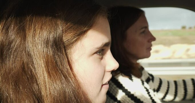 Close-up of daughter and mother in the background in a vehicle. The mother is driving and they are talking to each other. The sun is shining. The background is a clear field with grey clouds.