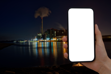 Cell phone with transparent screen in human hands. In the background, blurred, colorfully lit, a coal-fired power station with a smoking chimney.