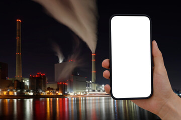 Cell phone with transparent screen in human hands. In the background, blurred, colorfully lit, a coal-fired power station with a smoking chimney.