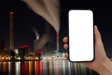Cell phone with transparent screen in human hands. In the background, blurred, colorfully lit, a coal-fired power station with a smoking chimney.