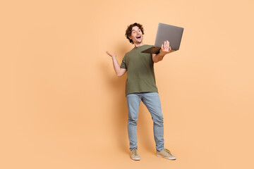 Young curly haired man gesturing happily while holding a laptop against a beige background in a trendy khaki t-shirt
