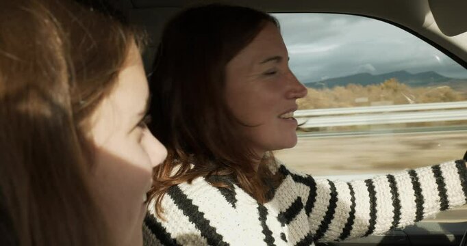 A daughter and her mother in a vehicle. The mother drives and looks at her daughter with complicity and smiles. The sun is shining. The background is a field with grey clouds. Focus on the mother.