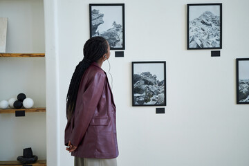 Person with long braids observing black-and-white landscape photographs on gallery wall while standing next to shelves with pottery and decor items