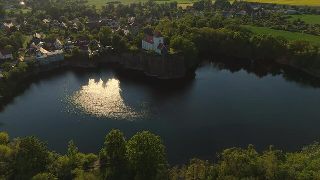 Drone footage of the Wehrkirche Beucha on a cliff above a heart-shaped quarry lake near Leipzig. Golden hour light highlights the unique shape, church, and surrounding village.
