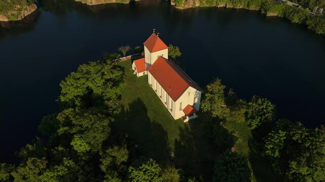 Drone view of the historic Wehrkirche Beucha in Saxony, Germany. The church stands atop a cliff surrounded by a flooded quarry, with the evening sun casting warm light across the landscape.