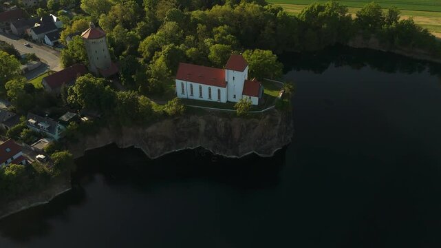 Aerial drone shot of the Wehrkirche Beucha in Saxony, perched on a rocky cliff above a quarry lake. Captures the historic church and surrounding forest in warm sunset light.