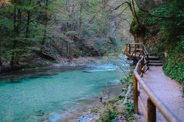 Scenic forest trail with a wooden railing path winding along a crystal-clear turquoise river, surrounded by lush green trees and rocky terrain