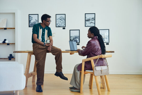 African American couple collaborating on work project in modern art gallery. Seated woman using laptop while man standing beside table, discussing project details