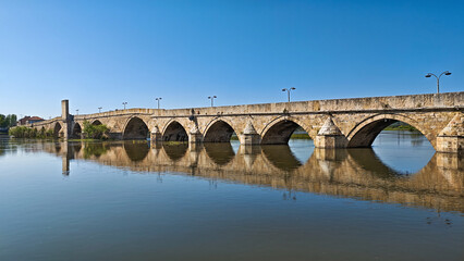 Panoramic image of the historic Old Bridge over the Maritsa River in Svilengrad, Bulgaria, reflecting beautifully in the calm blue water under a clear sky.