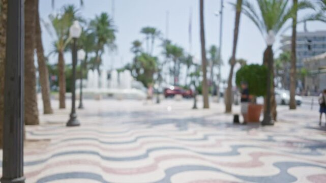 Outdoor blurred view of alicante rambla with defocused palm trees fountain and walking people during a sunny day in spain