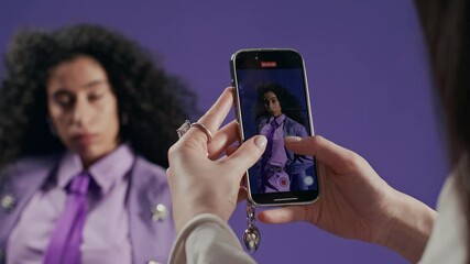 Cropped shot of hands of unrecognizable woman holding smartphone recording video of beautiful biracial female model in stylish lilac suit posing in professional studio