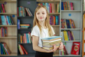 Young girl in a library holds multiple books while smiling confidently. Back to school.