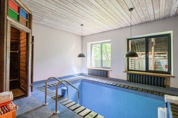 Indoor pool room with blue tile, a wooden sauna entrance, and two windows. Modern light fixtures and a whitewashed wood ceiling complete the scene