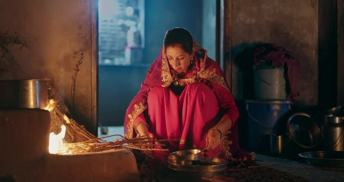Indian villager gen x woman preparing food using traditional earthen mud stove indoor house asian young female wear red saree cooking on open fire flam inside kitchen home