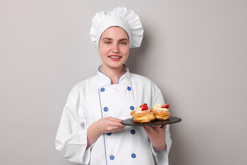 Confectioner in uniform holding delicious profiteroles on light grey background